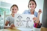 Portrait of mother and daughter sitting at table and daughter showing her drawing-1