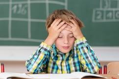 Young boy concentrating on his schoolwork sitting at his desk in the classroom with his head in his hands reading his class notes.jpeg