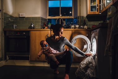 Young mother with children with autism is holding her crying baby daughter on her hip while trying to load the washing machine