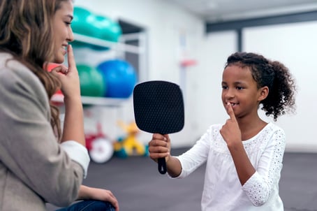 child with autism holding mirror and touching lips for therapy exercises with the guidance of an adult female therapist